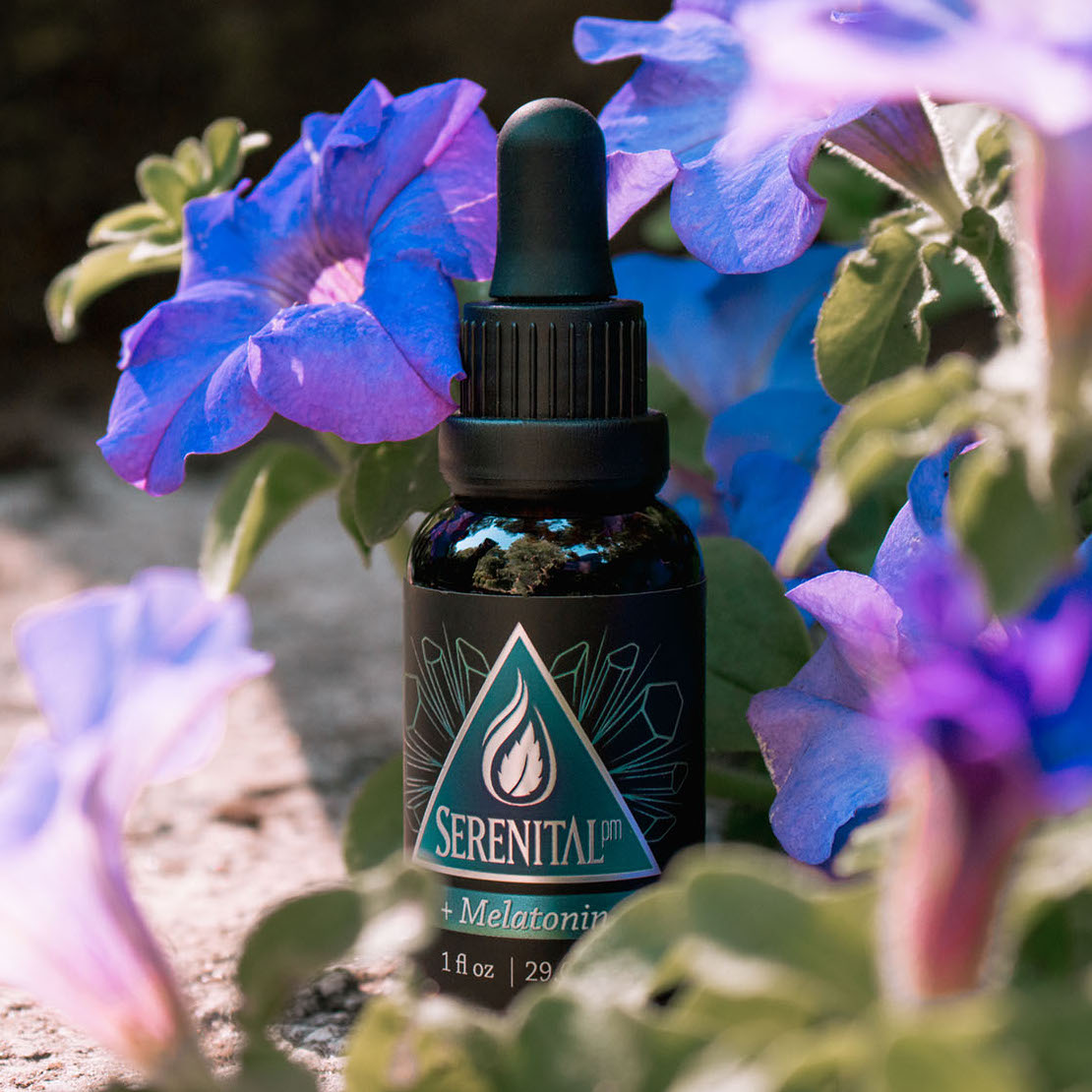 A bottle of Serenital CBD + Melatonin sits on a rock amongst some deep purple and blue colored flowers. The flowers appear to be morning glory.